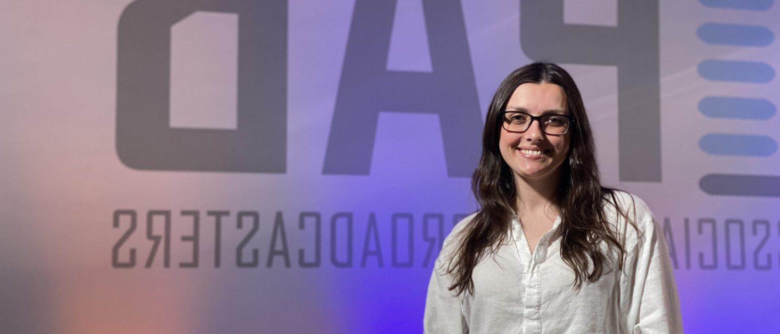 Image shows Makenzie Christman holding her PAB Award in front of a wall that says, 